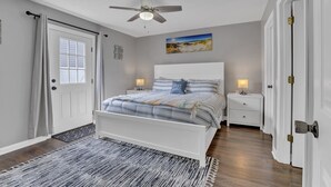 A bedroom with a white bed, a ceiling fan, a nightstand with a lamp, and a door with a doorknob.