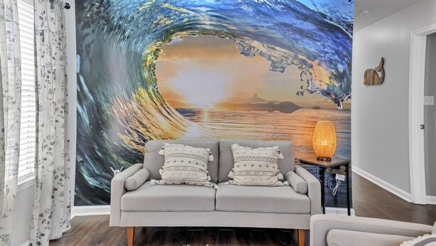 A room with a large wave mural on the wall, a grey sofa with patterned cushions, a small table with a lamp, and a wooden floor.