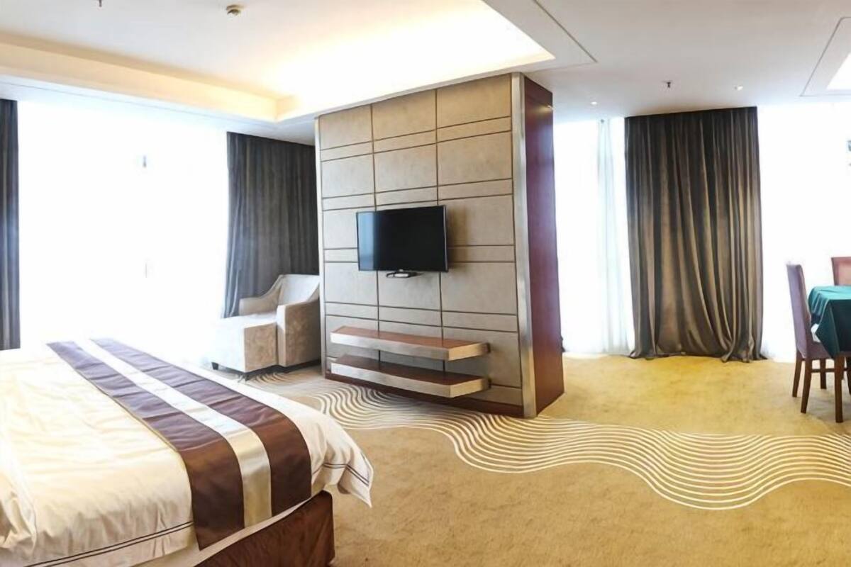 A hotel room with a large bed, a TV mounted on a wall, a seating area, and a view of the outdoors.
