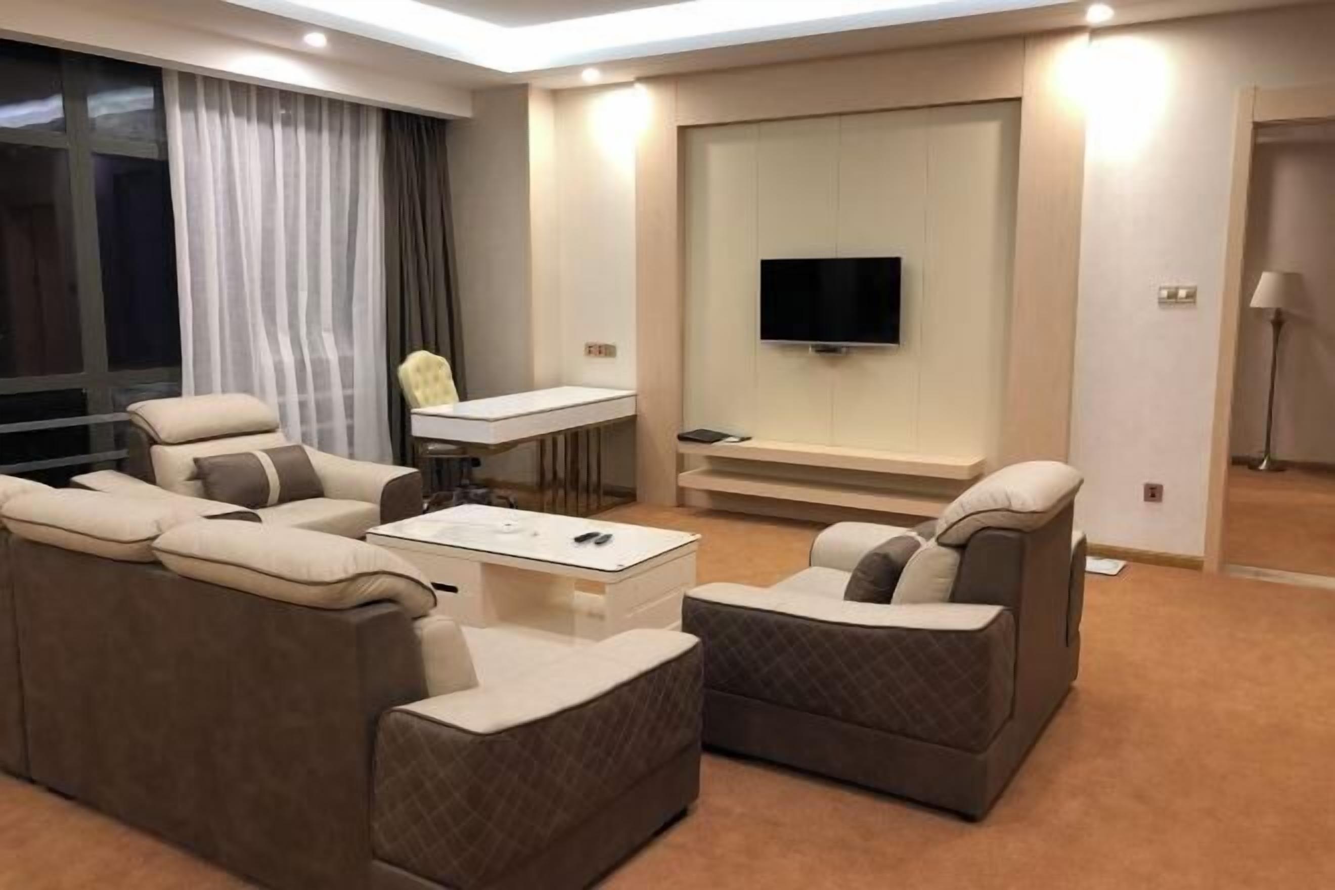 A modern hotel room with a sofa set, a coffee table, a TV mounted on the wall, and a desk with a chair.