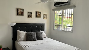 A bedroom with a black headboard, white bedding, and a window with a view of greenery and parked cars. - acogedor apartamento acceso directo al jardín en primera linea zona con segurida (La Guáyiga)