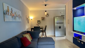 A modern living room with a grey sofa, a dining area with a table and chairs, a built-in TV, and a stainless steel refrigerator. - acogedor apartamento acceso directo al jardín en primera linea zona con segurida (La Guáyiga)