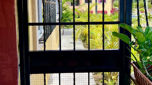 A view through a barred window with a metal gate, overlooking a street with buildings and greenery. - acogedor apartamento acceso directo al jardín en primera linea zona con segurida (La Guáyiga)