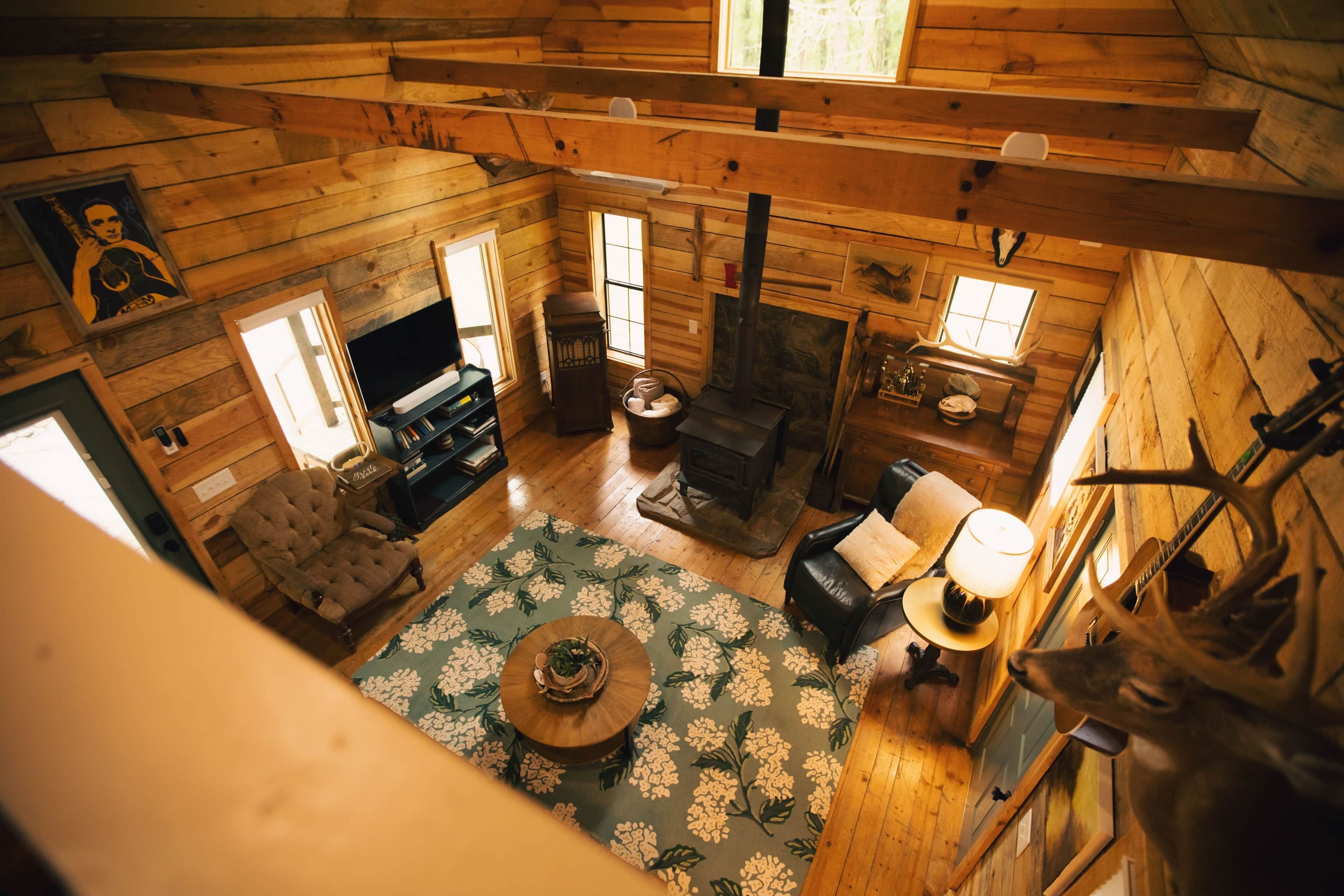 Interior de una cabaña con chimenea, paredes de madera, un sofá, una mesa de comedor y un mantel con estampado floral, además de un televisor.
