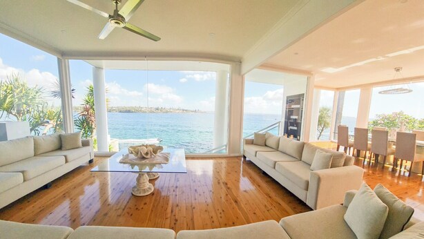 Un salón amplio con suelo de madera, dos sofás color beige, una mesa de centro de vidrio y vista al mar.