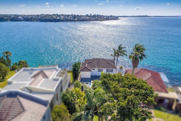 A coastal view with a house overlooking the sea, surrounded by greenery and a clear sky. - Waterfront Paradise in Royal National Park - Your Bundeena Escape (Bundeena)