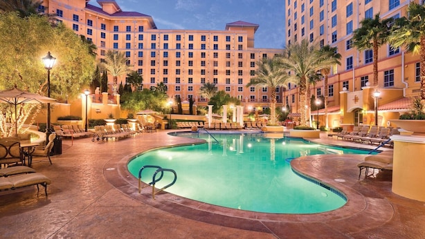 Ein Hotelpoolbereich mit Liegestühlen und Sonnenschirmen, umgeben von Palmen und einem großen Gebäude im Hintergrund.