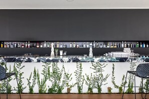 A bar area with a long shelf of liquor bottles, a white wall with green plants, and two black chairs.