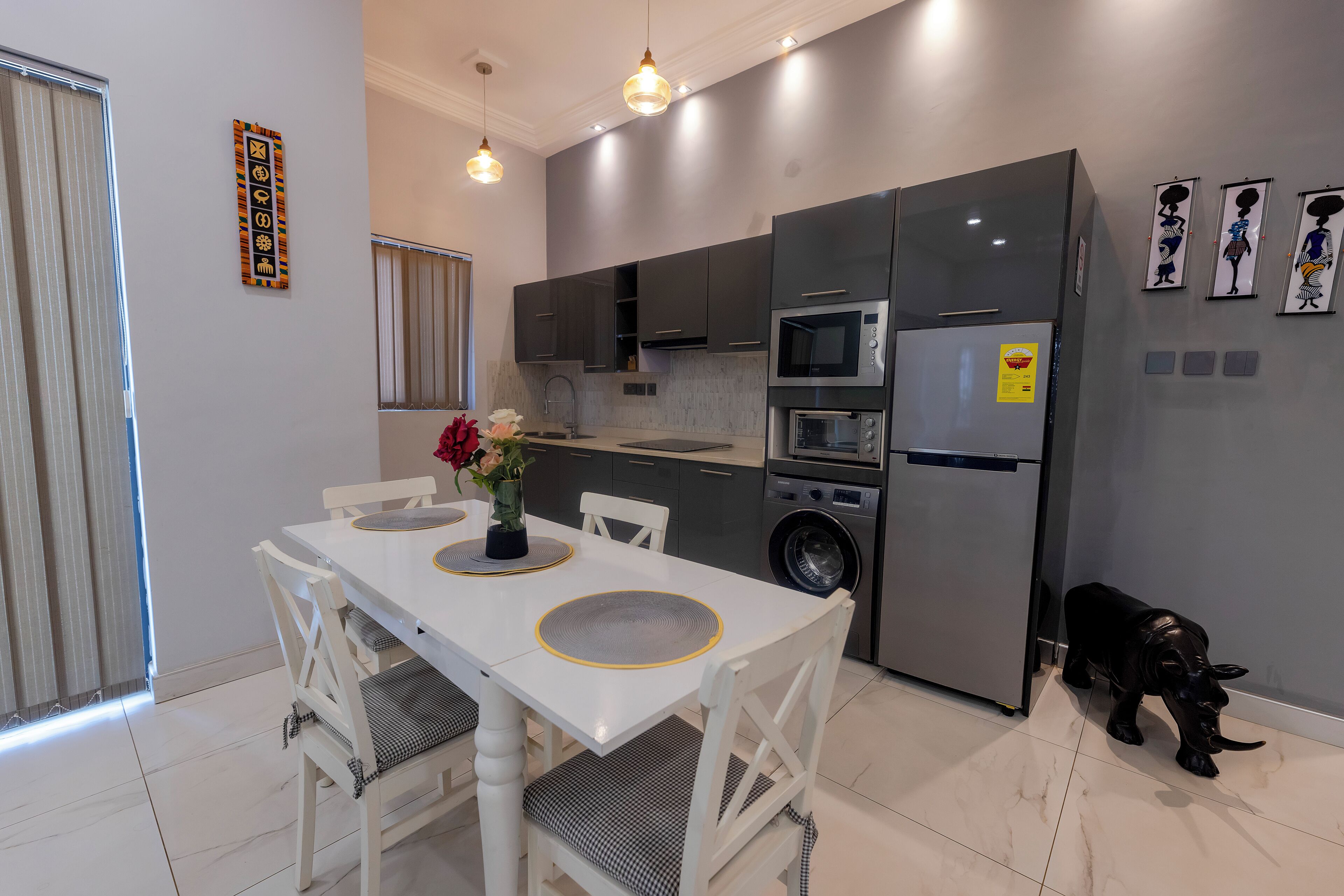 A modern kitchen with grey cabinets, a stainless steel refrigerator, and a washing machine. There is a dining table with chairs and a vase of flowers.