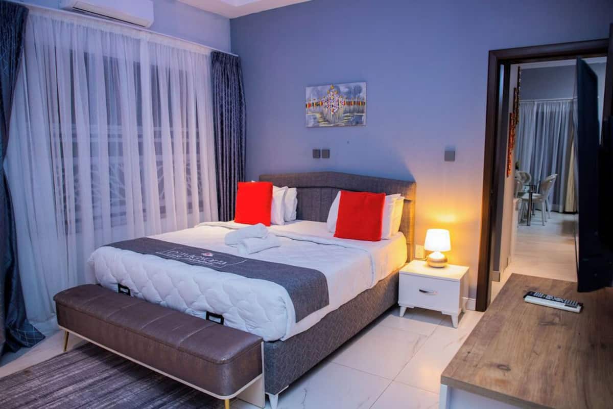 A modern hotel room with a large bed, a bedside table with a lamp, a bench, and a wooden side table.