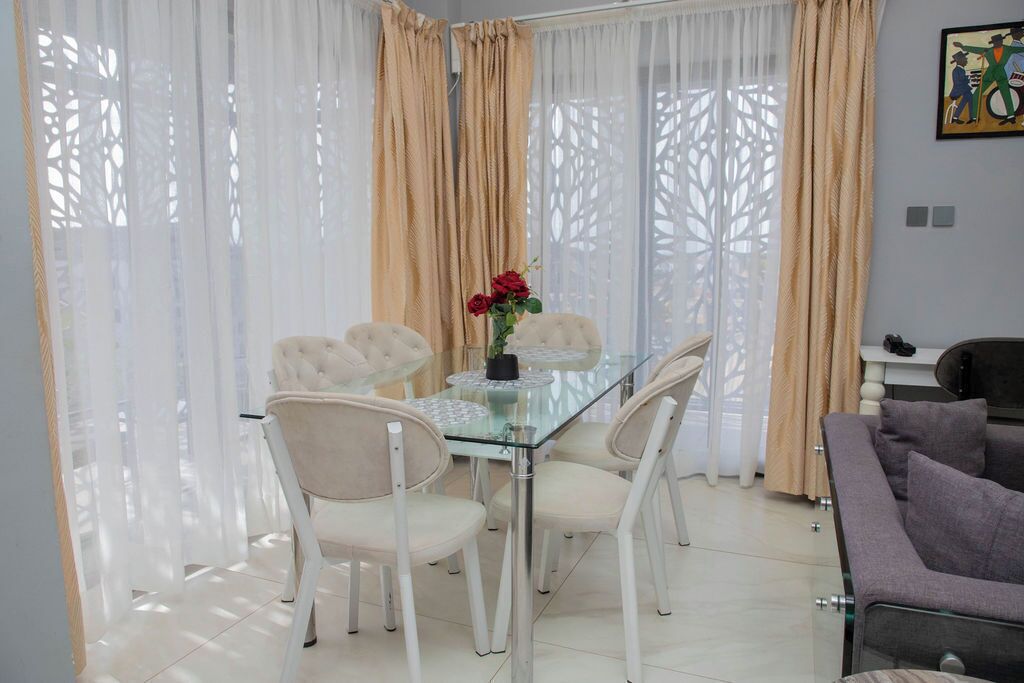 A dining area with a glass table and white chairs, beige curtains, and a vase with red flowers.