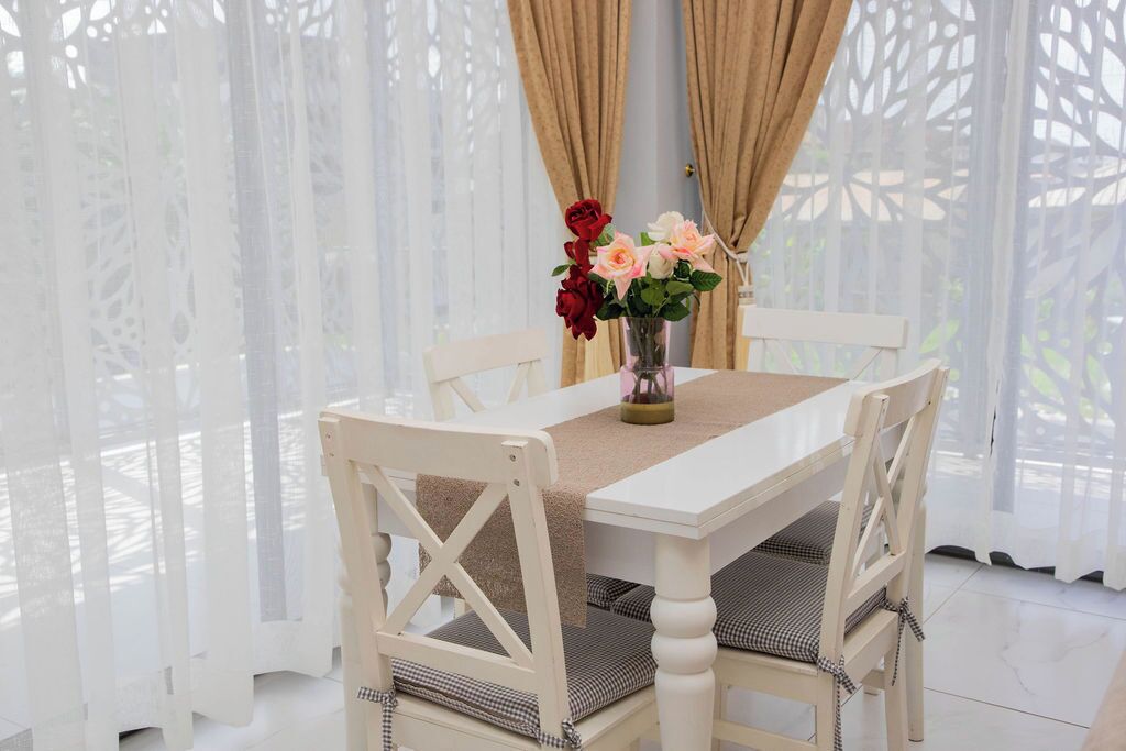 A white dining table with a beige table runner, surrounded by white chairs with checkered cushions. A vase with flowers is placed on the table.