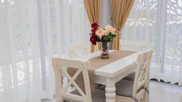 A white dining table with a beige table runner, surrounded by white chairs with checkered cushions. A vase with flowers is placed on the table.