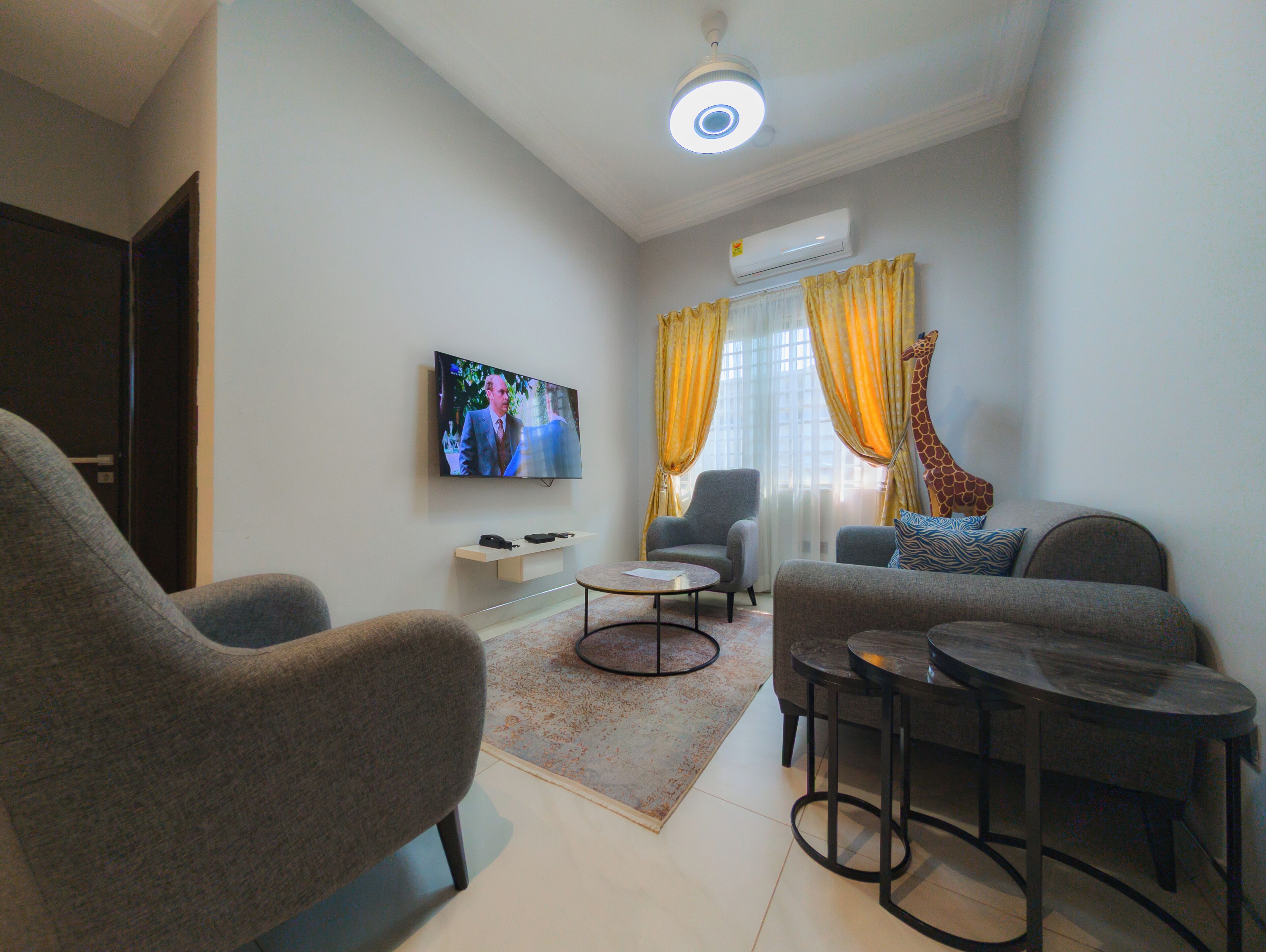 A living room with a flat-screen TV, grey sofas, a coffee table, and a ceiling light fixture.
