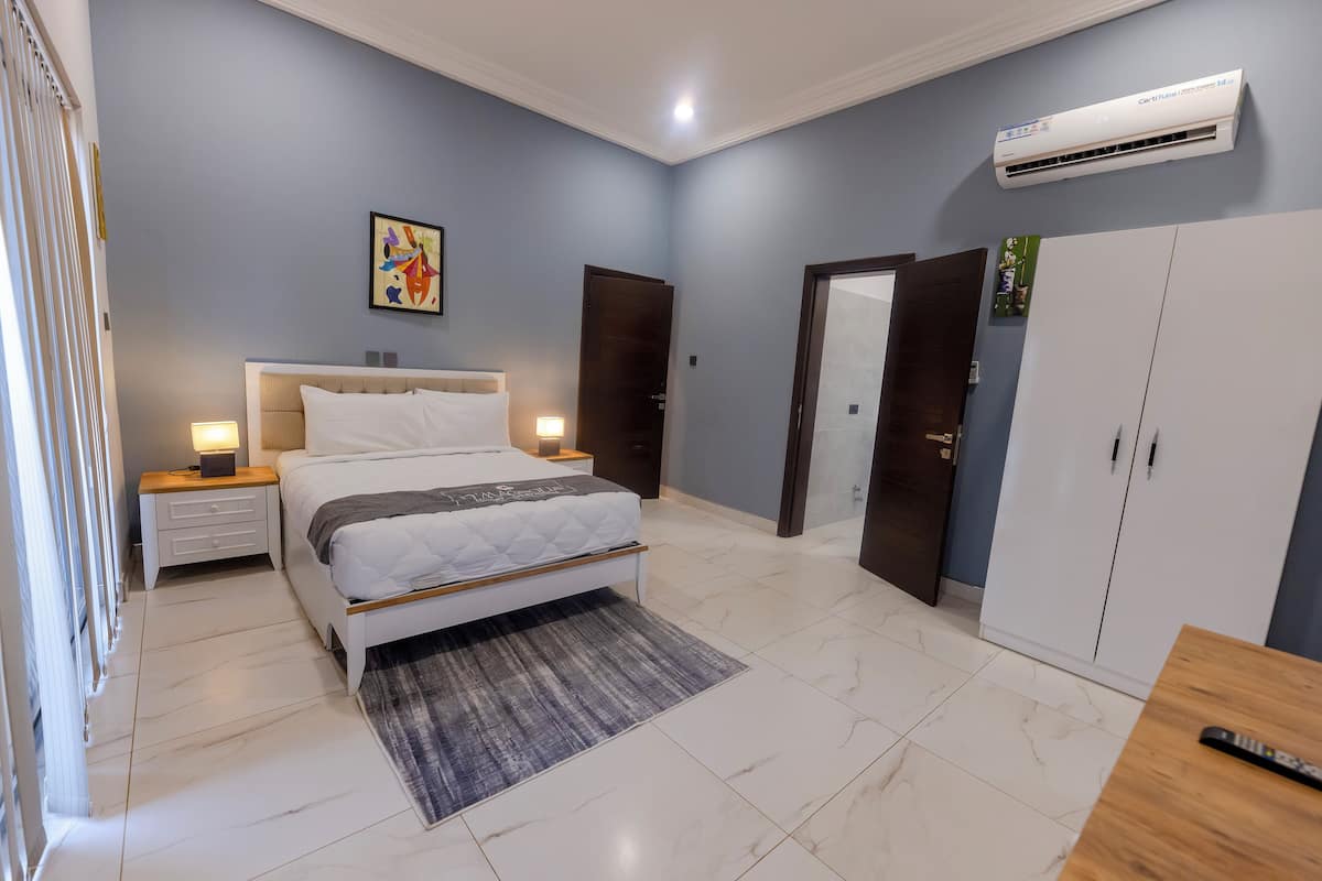 A bedroom with a bed, bedside tables, a nightstand, a wardrobe, and an air conditioning unit.