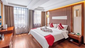 Een hotelkamer met een groot bed, houten vloeren, een bureau en uitzicht op de stad door grote ramen.
