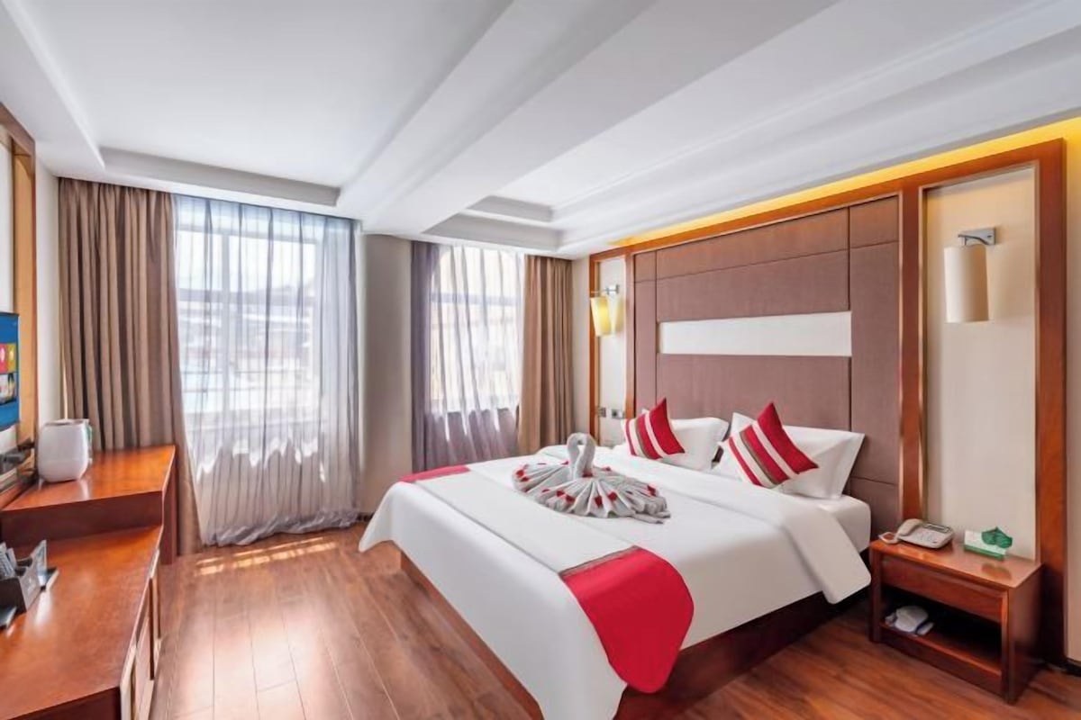 Habitación de hotel con una cama grande, suelo de madera, un escritorio y vistas de la ciudad a través de amplias ventanas.