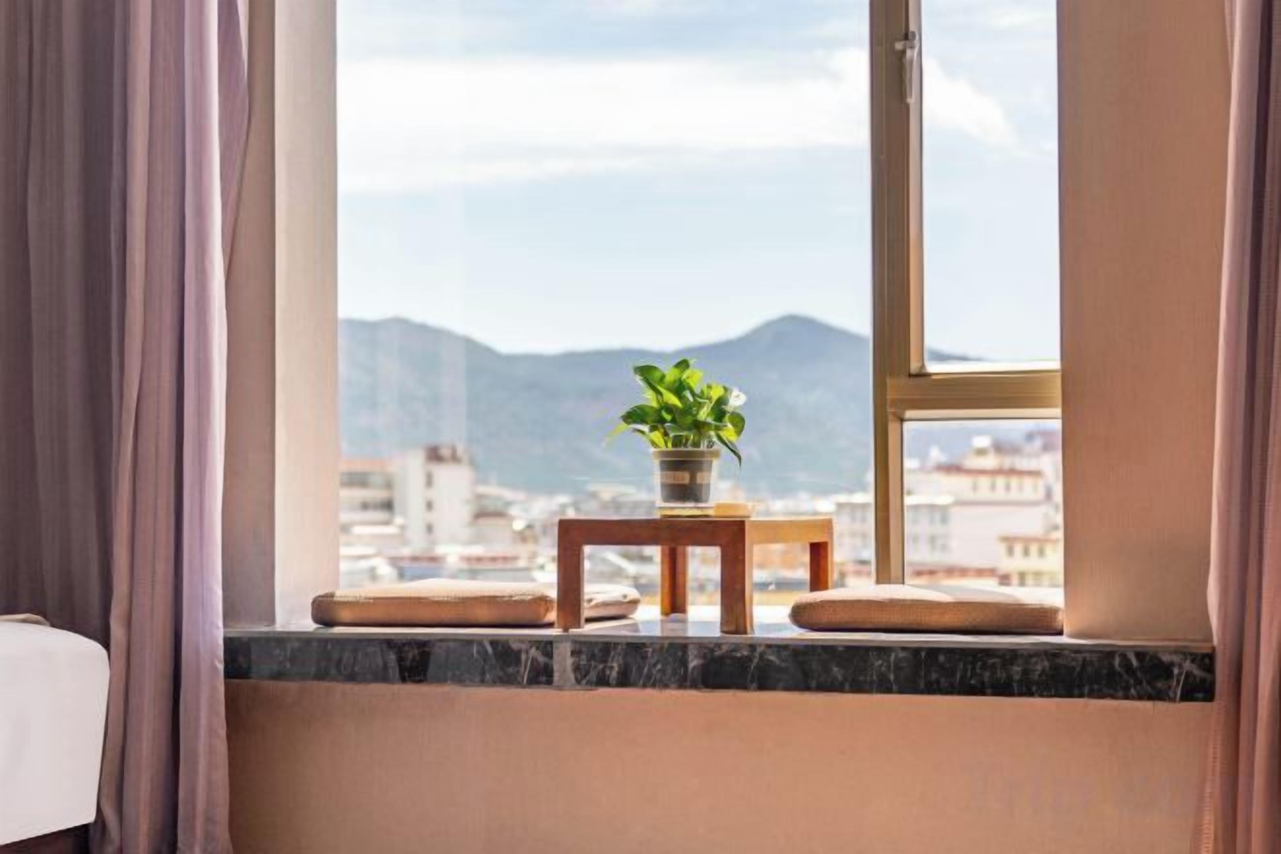 A window with a view of mountains and a cityscape, a small table with a plant, and a cushion on the windowsill.