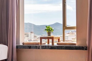 Janela com vista para montanhas e para a cidade, uma mesinha com uma planta e um almofada no parapeito.