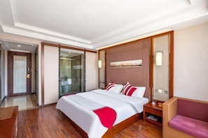 Habitación de hotel con una cama grande, piso de madera, puerta de vidrio que da al baño y una lámpara fijada a la pared.