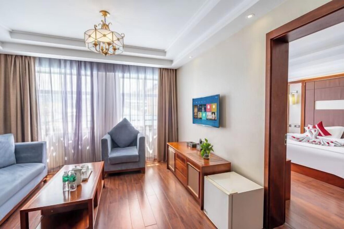 A hotel room with a wooden floor, a grey sofa, a grey armchair, a wooden side table with a plant, and a bed with white bedding.