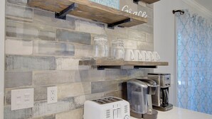 A kitchen with a rustic wooden backsplash, a white countertop, a toaster, a coffee maker, and a shelf with jars and cups. - Steps to Oak Island Beach | Private Pool | Pet Friendly | Beach Gear (Oak Island)