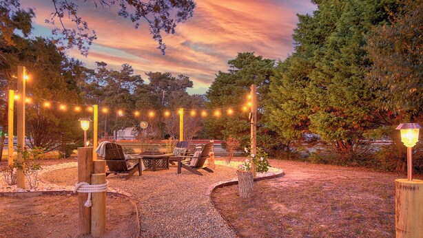 An outdoor seating area with a table and chairs, surrounded by a gravel path and string lights. - Steps to Oak Island Beach | Private Pool | Pet Friendly | Beach Gear (Oak Island)