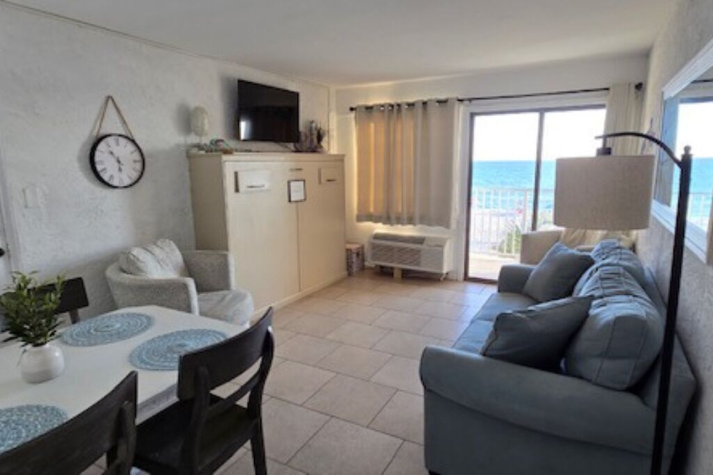 Un salon moderne comprenant un canapé, une table à manger avec des chaises, un téléviseur posé sur un meuble et une vue sur la mer grâce à de grandes fenêtres.