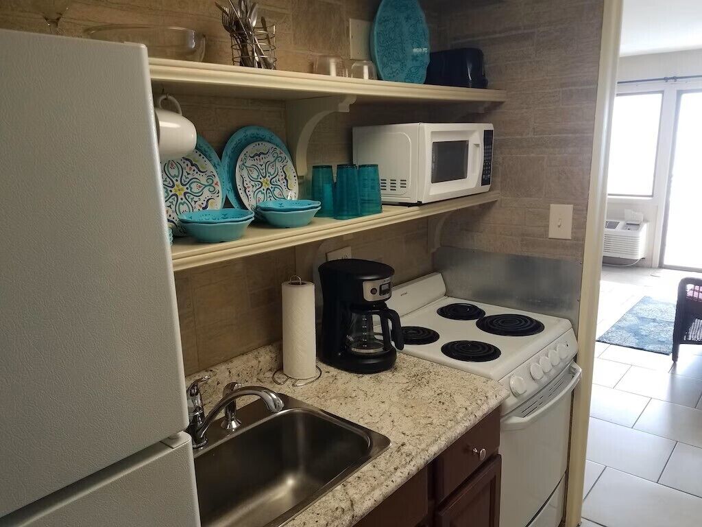 Una cocina con un refrigerador blanco, una estufa, un microondas y un lavamanos. Hay vajilla azul y blanca en los estantes.