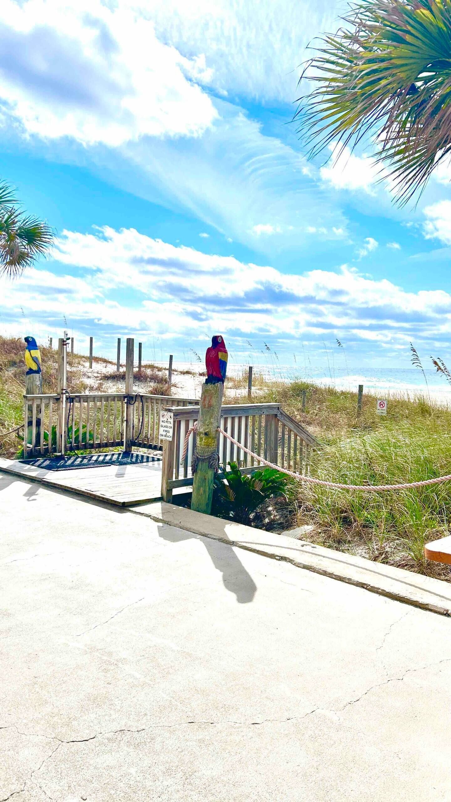 A wooden boardwalk with a metal railing, leading to a beach area with sand and sparse vegetation.