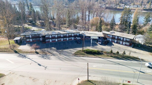 A multi-story building with a parking lot in front, surrounded by trees and a body of water in the background. - GOLDPAN MOTEL  (Quesnel)