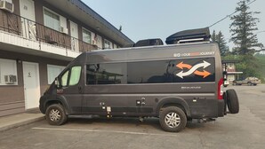 A gray van parked in front of a building with numbered doors, a balcony with a railing, and a clear sky. - GOLDPAN MOTEL  (Quesnel)