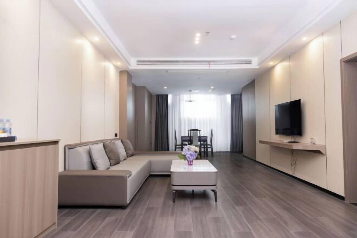 A modern hotel room with a sectional sofa, a small coffee table, a flat-screen TV, and a dining area with a table and chairs.