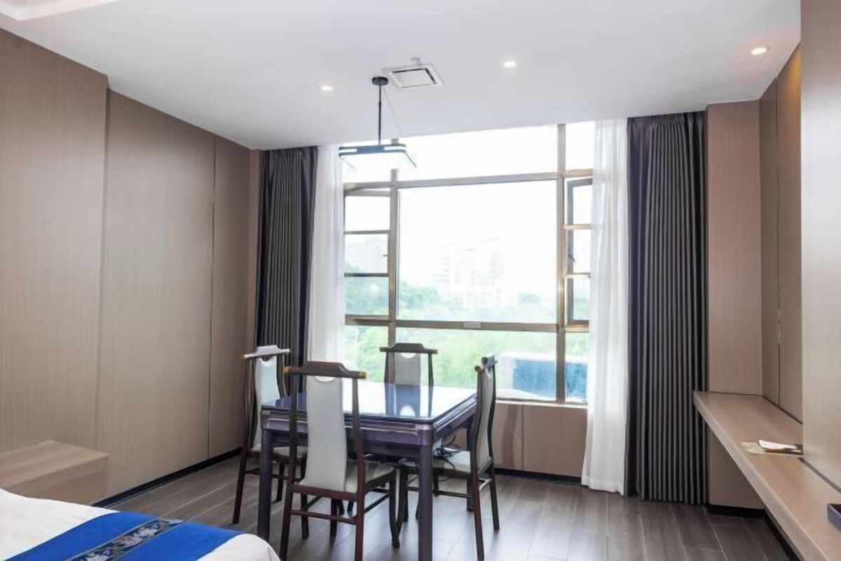A hotel room with a bed, a dining table with chairs, a large window with curtains, and a ceiling light fixture.