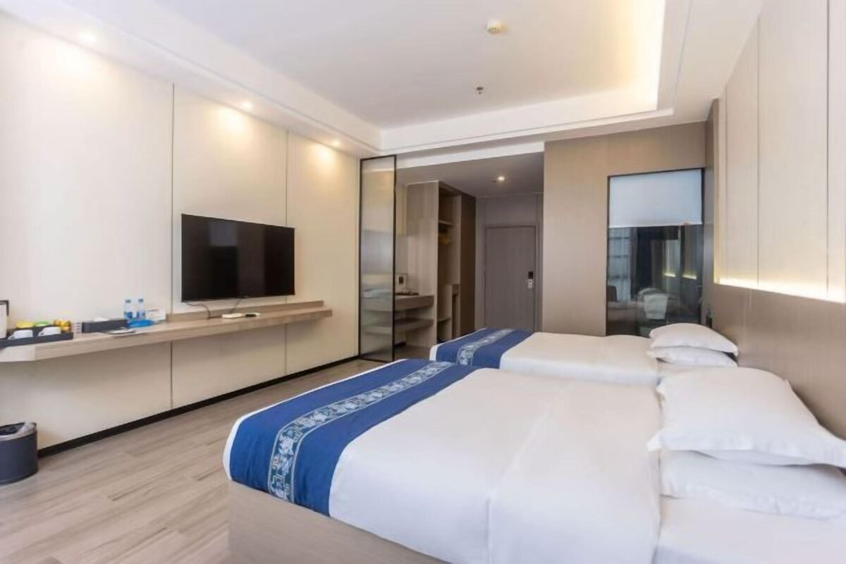 A hotel room with two beds, a TV, a mirror, and a bathroom visible through a glass door.