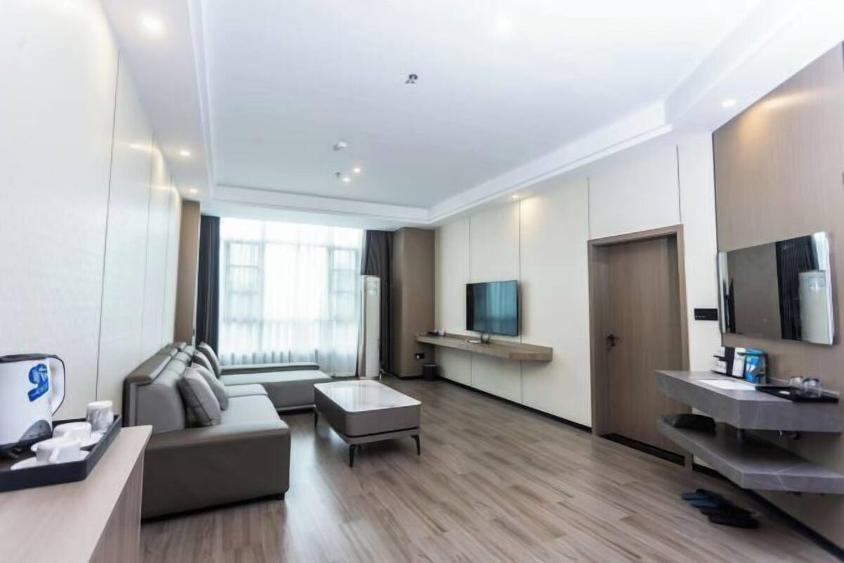 A modern hotel room with a sofa, a coffee table, a TV mounted on the wall, and a wooden floor.