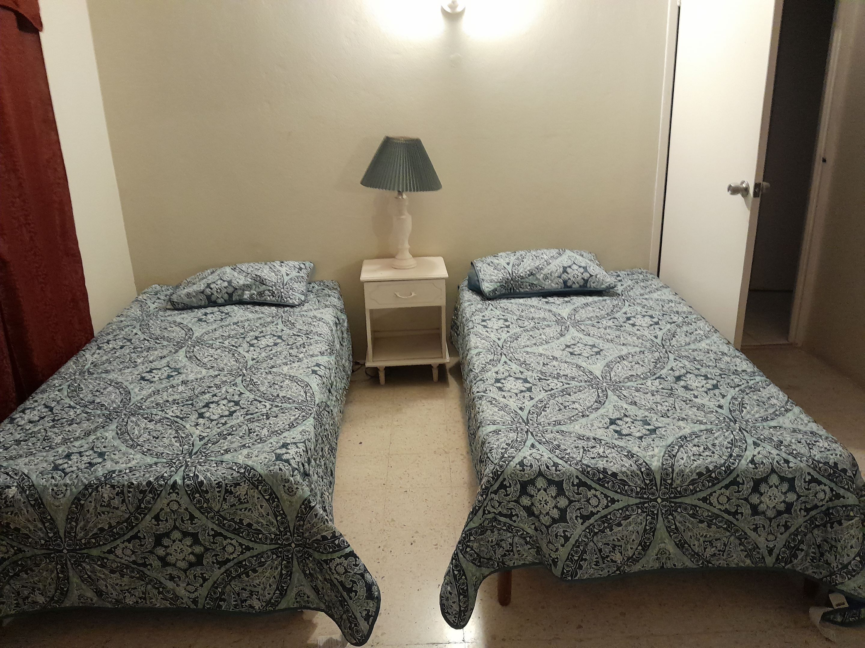 A room with two beds, each with a patterned bedspread, a bedside table with a lamp, and a door to the right.