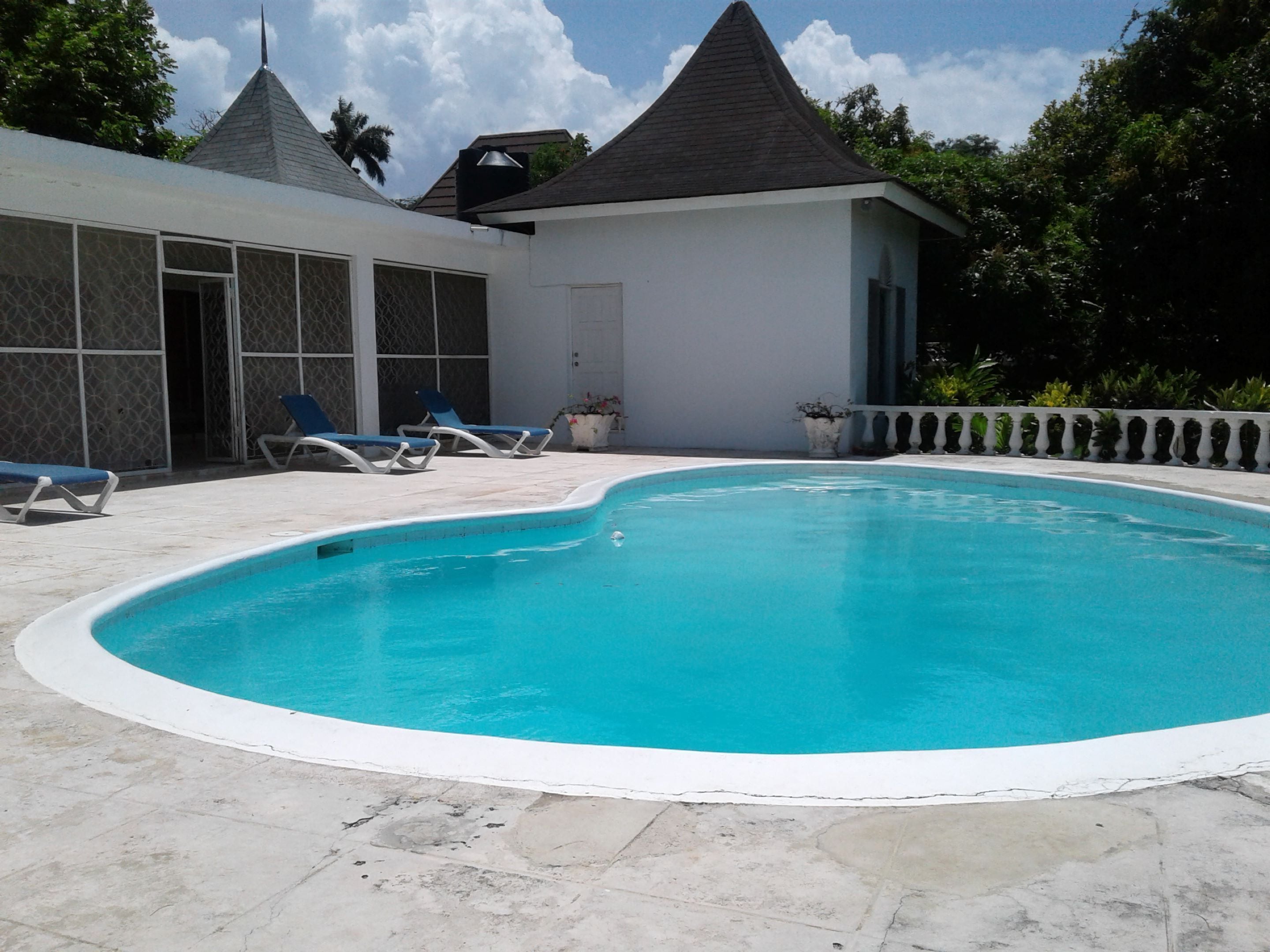A swimming pool with lounge chairs, a covered area with a peaked roof, and a white building with patterned windows.