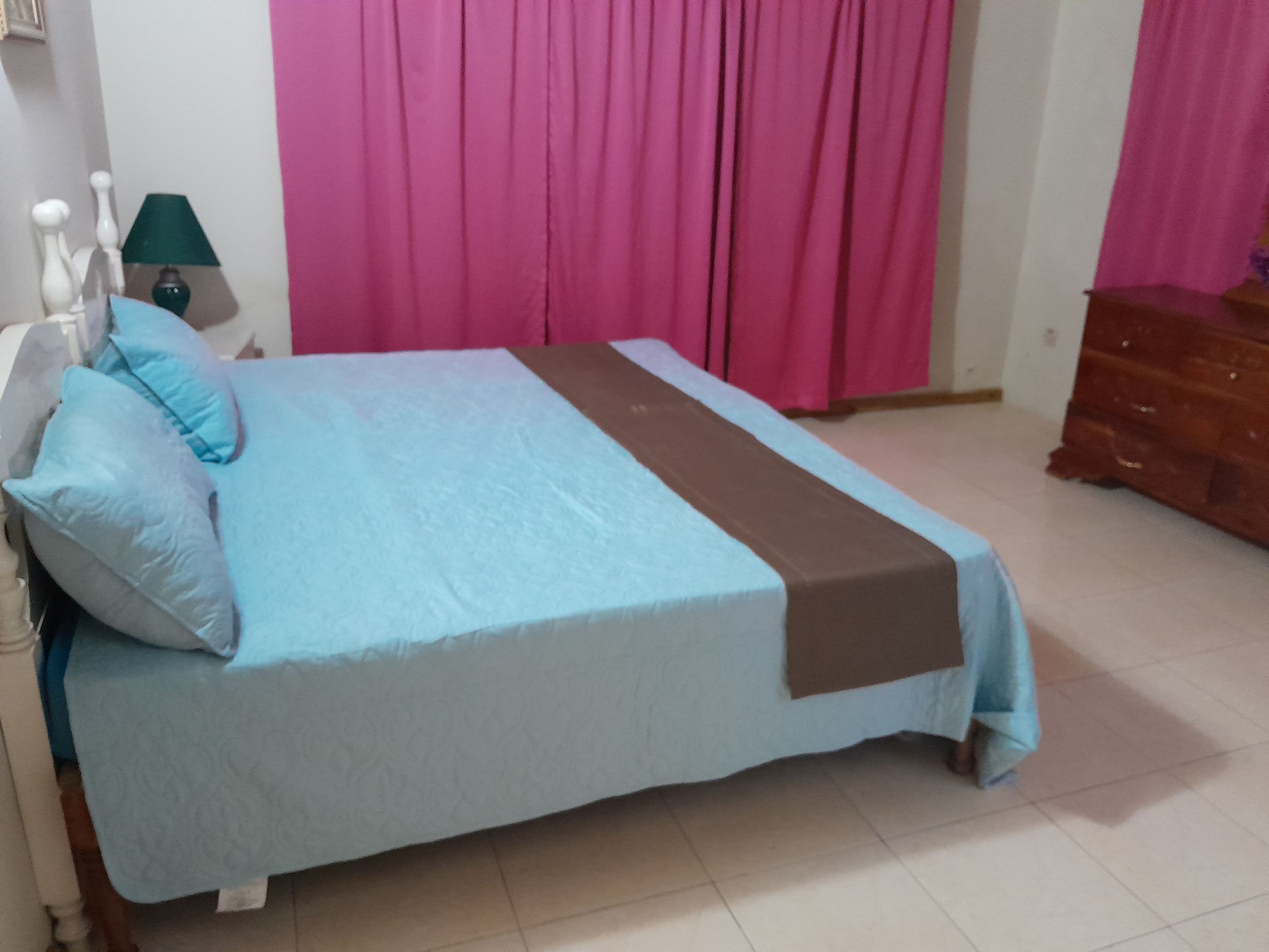 Una cama con una colcha azul y marrón, un aparador de madera y cortinas rosas.