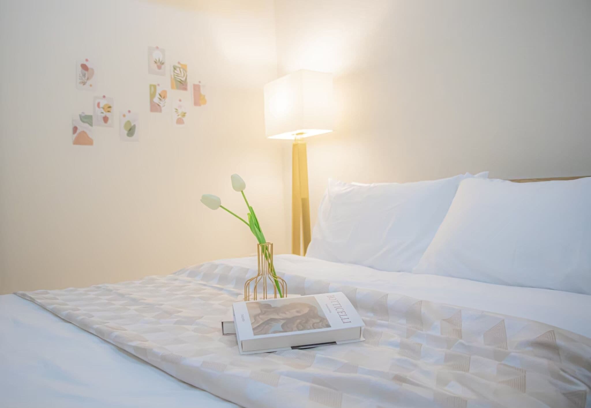 A neatly made bed with a book and a vase of flowers, a lamp, and wall art.