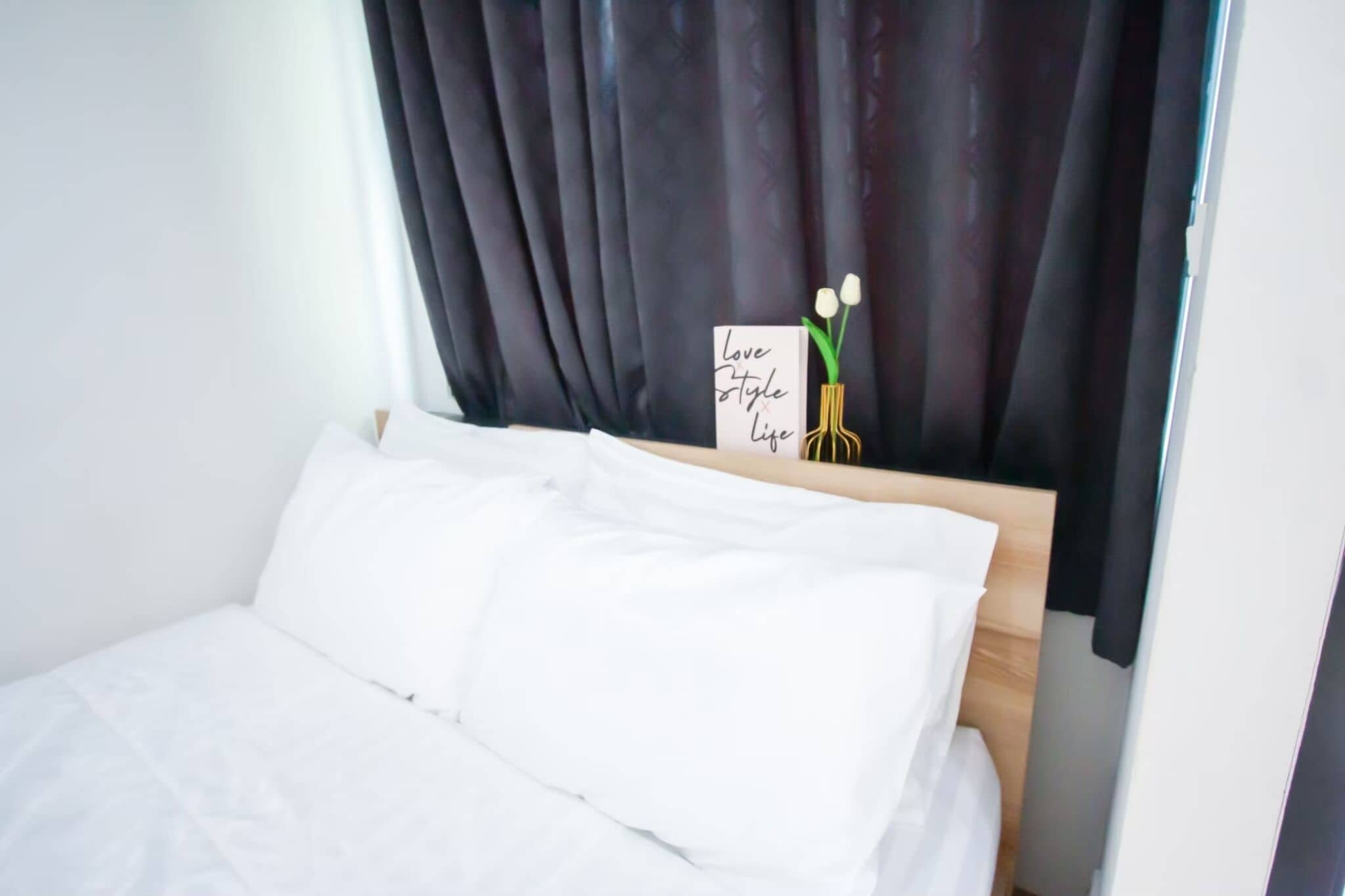 A neatly made bed with white linens, a dark curtain, and a sign with flowers and the words "Love Style Life".