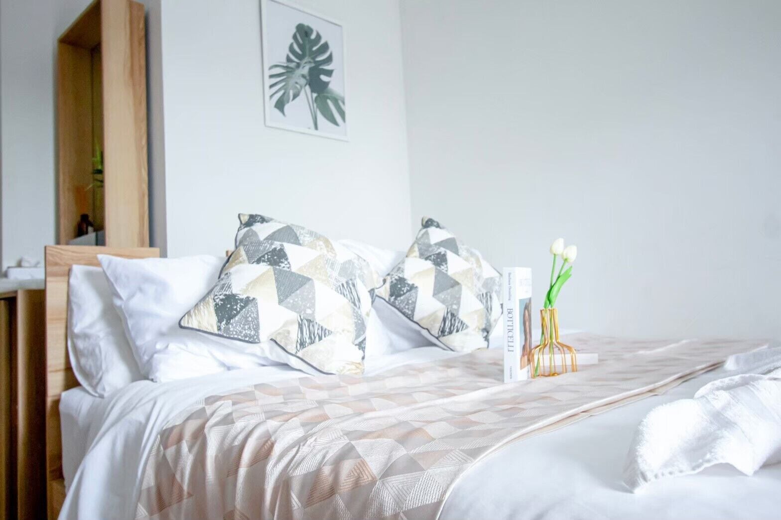 A neatly made bed with patterned pillows, a decorative vase with flowers, and a framed plant on the wall.