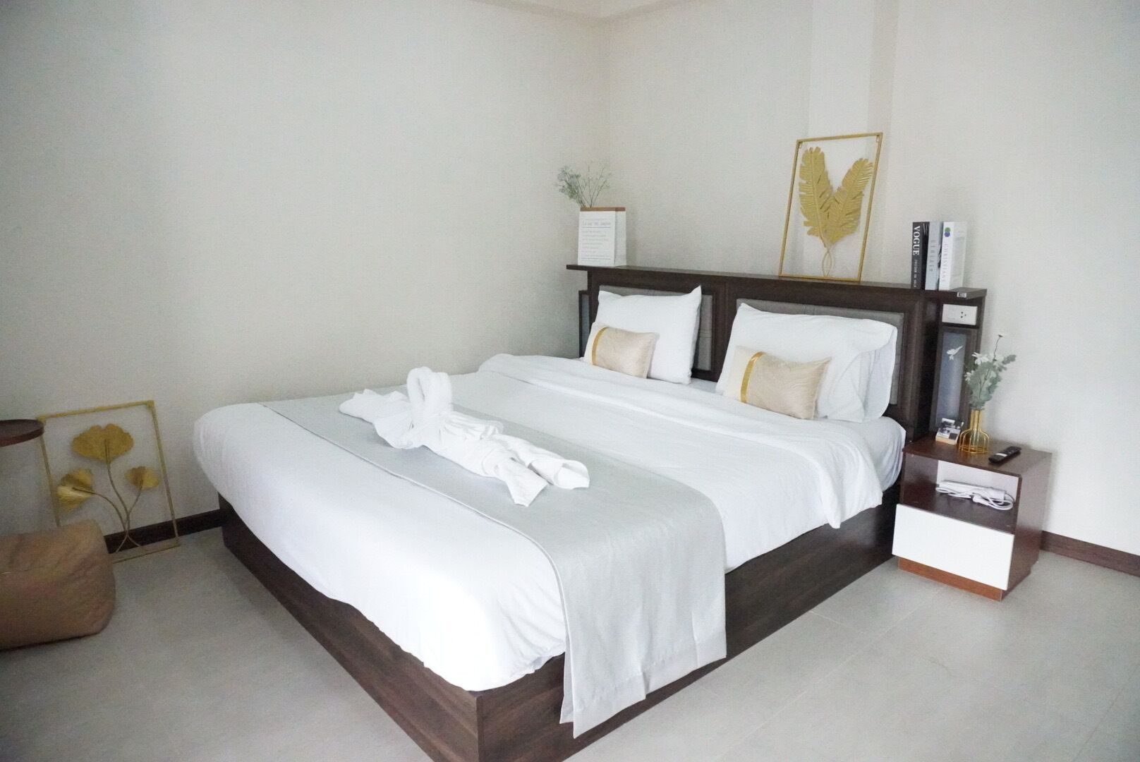 A neatly made bed with white linens and a towel, a bedside table with a vase and a remote control, and a framed artwork on the wall.