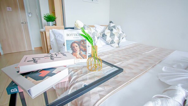 A neatly arranged hotel room with a bed, a glass table with books and a decorative vase, a framed picture on the wall, and a small potted plant. - ATC Residence (Bangkok)