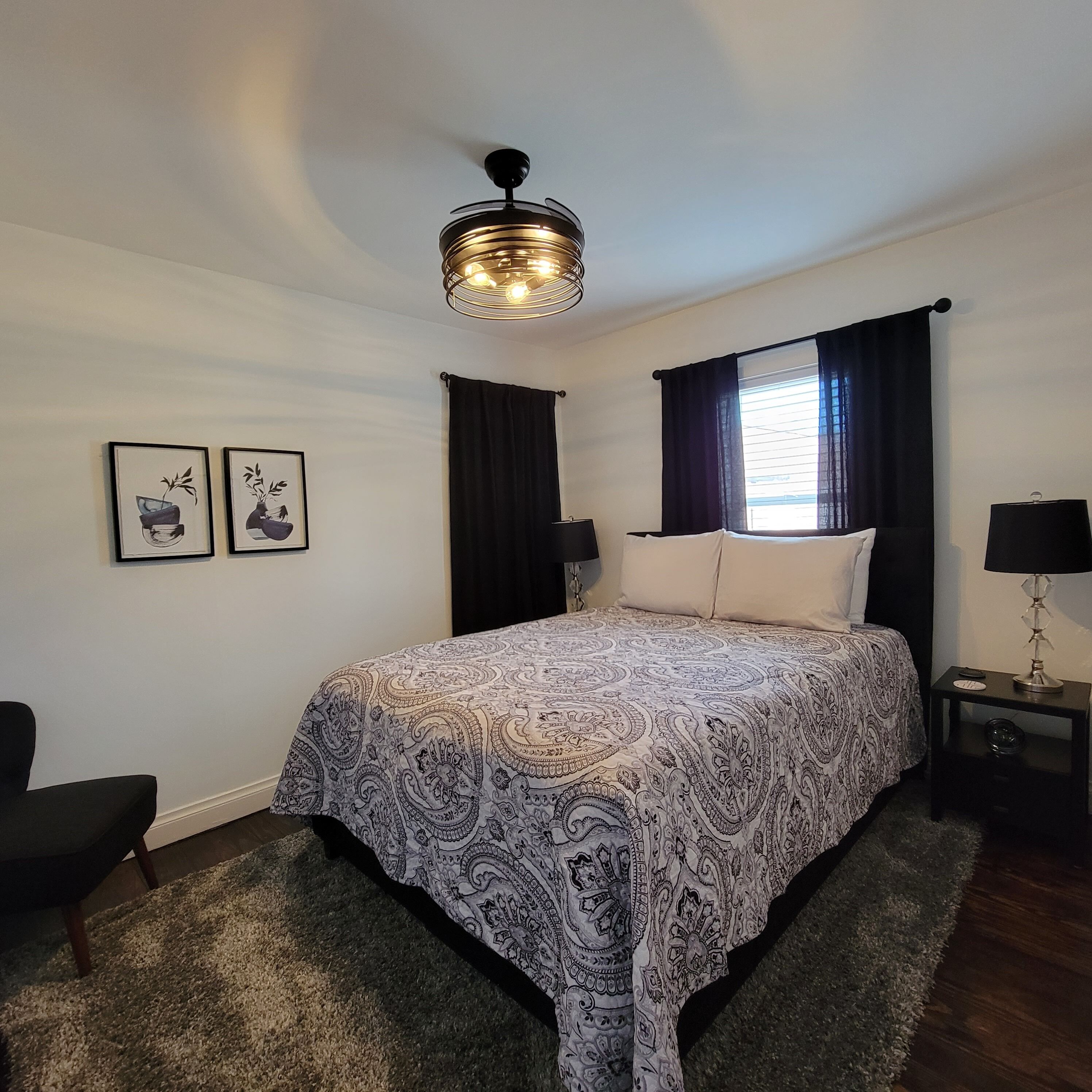 Un dormitorio con cama, ventilador de techo, dos lámparas de pared, ventana con cortinas y dos cuadros en la pared.