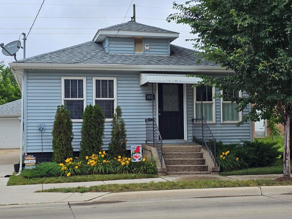 A two-story house with a front porch, a mailbox, and a small garden with yellow flowers. - Close to the lake, pet friendly, fenced yard, nicely redone inside. Sleeps eight (Muskegon)