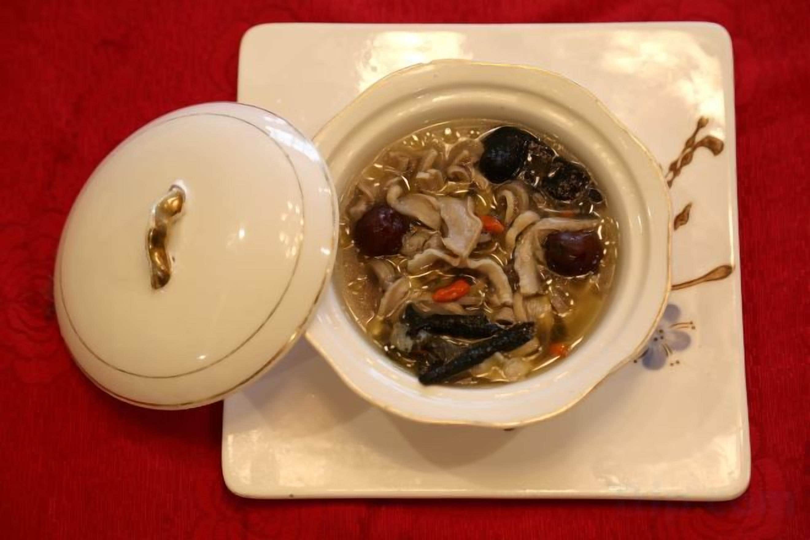 A bowl of soup with a lid on a white plate, placed on a red surface.