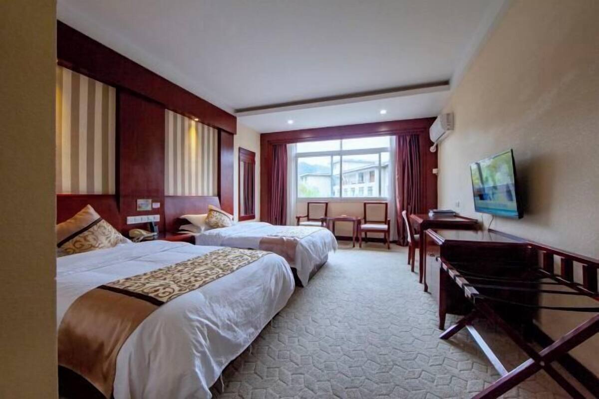 A hotel room with two beds, a desk, a chair, a TV, and a window with a view of buildings.