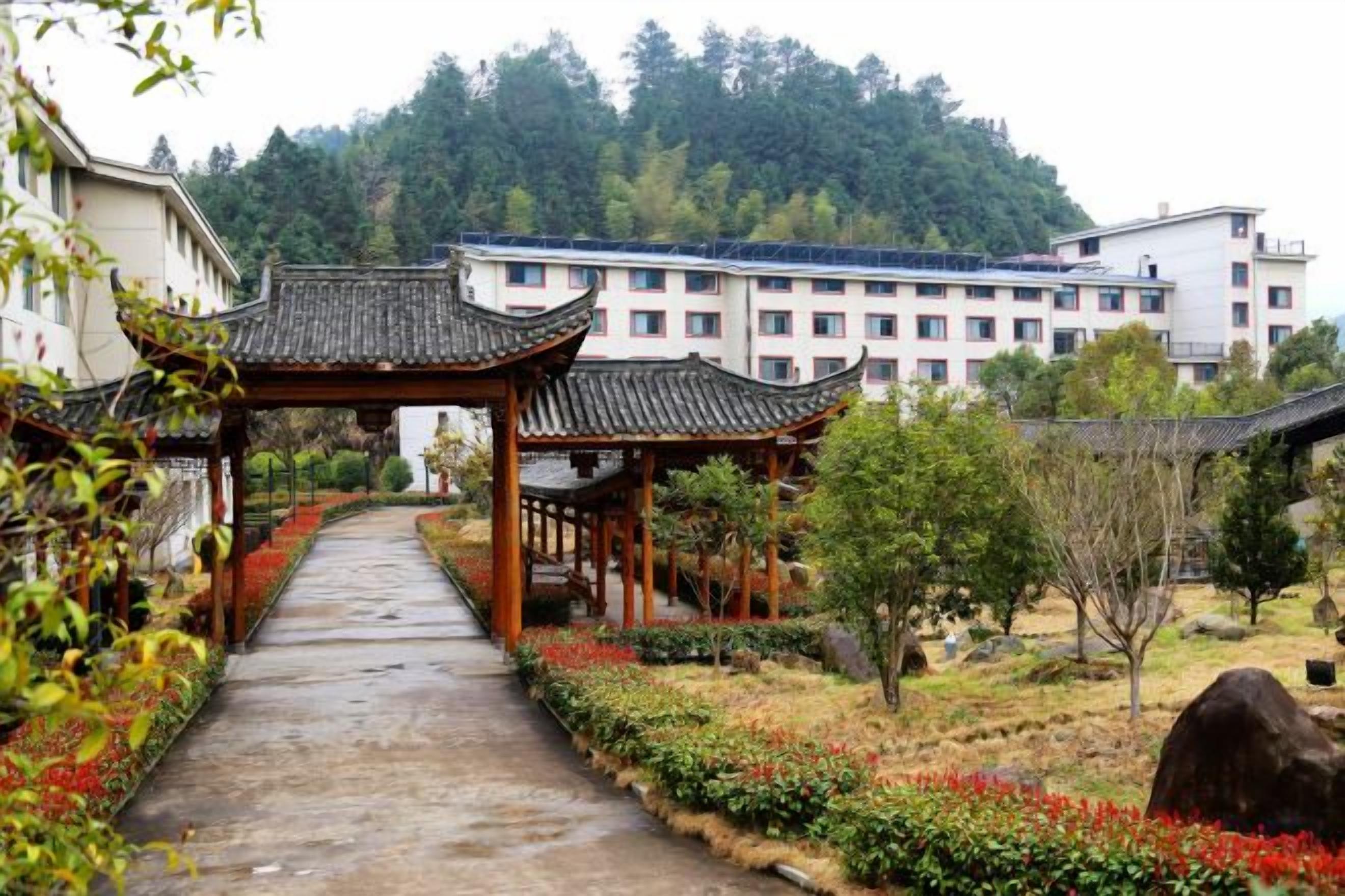 A pathway leading to traditional-style buildings with a view of a forested hill in the background.