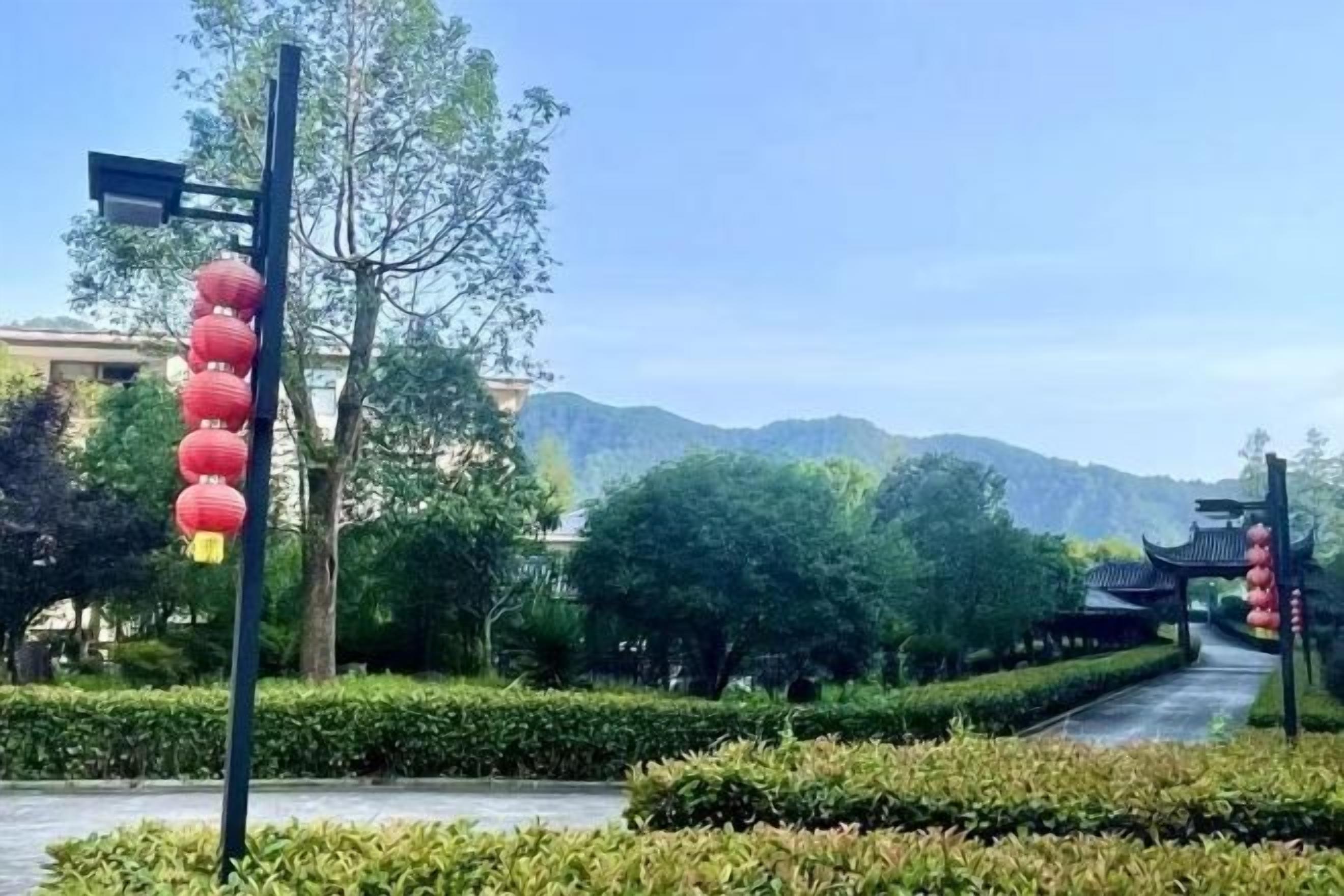 A pathway leading to a traditional gate with red lanterns, surrounded by greenery and a mountain backdrop.